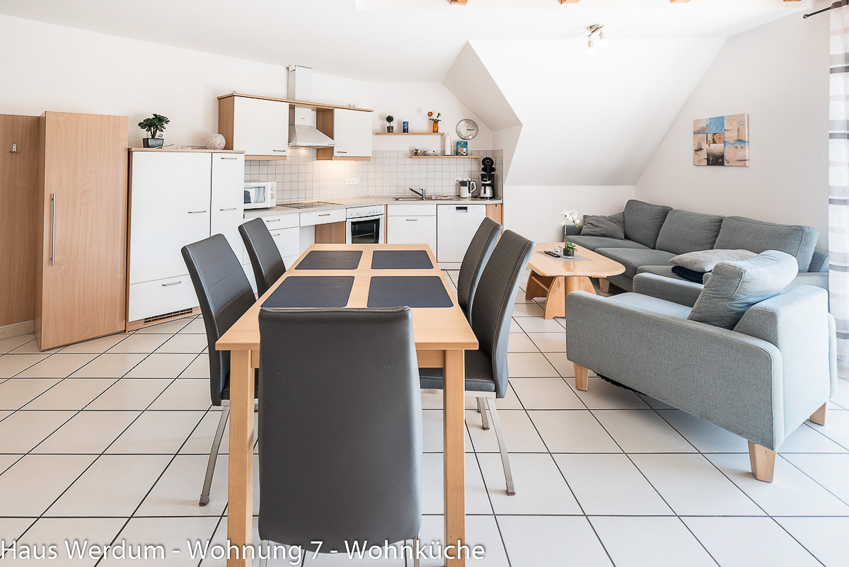 Großer Essplatz in der offenen Wohnküche der Wohnung 7 mit sechs Stühlen, Küchenzeile und angrenzendem Wohnbereich. Der unterfahrbare Esstisch liegt in einem Wohnraum von etwa 6 × 6 m mit 160 cm schmalstem Durchgang.