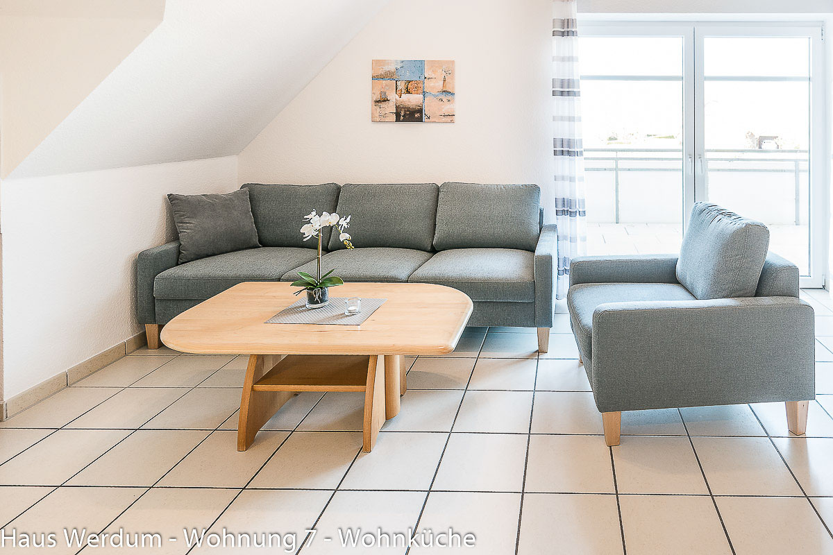 Wohnbereich der Wohnung 7 mit Sofa, Sessel, Couchtisch und direktem Balkonzugang auf durchgehend gefliestem Boden. Der Wohnraum misst etwa 6 × 6 m; der schmalste geprüfte Durchgang beträgt 160 cm.