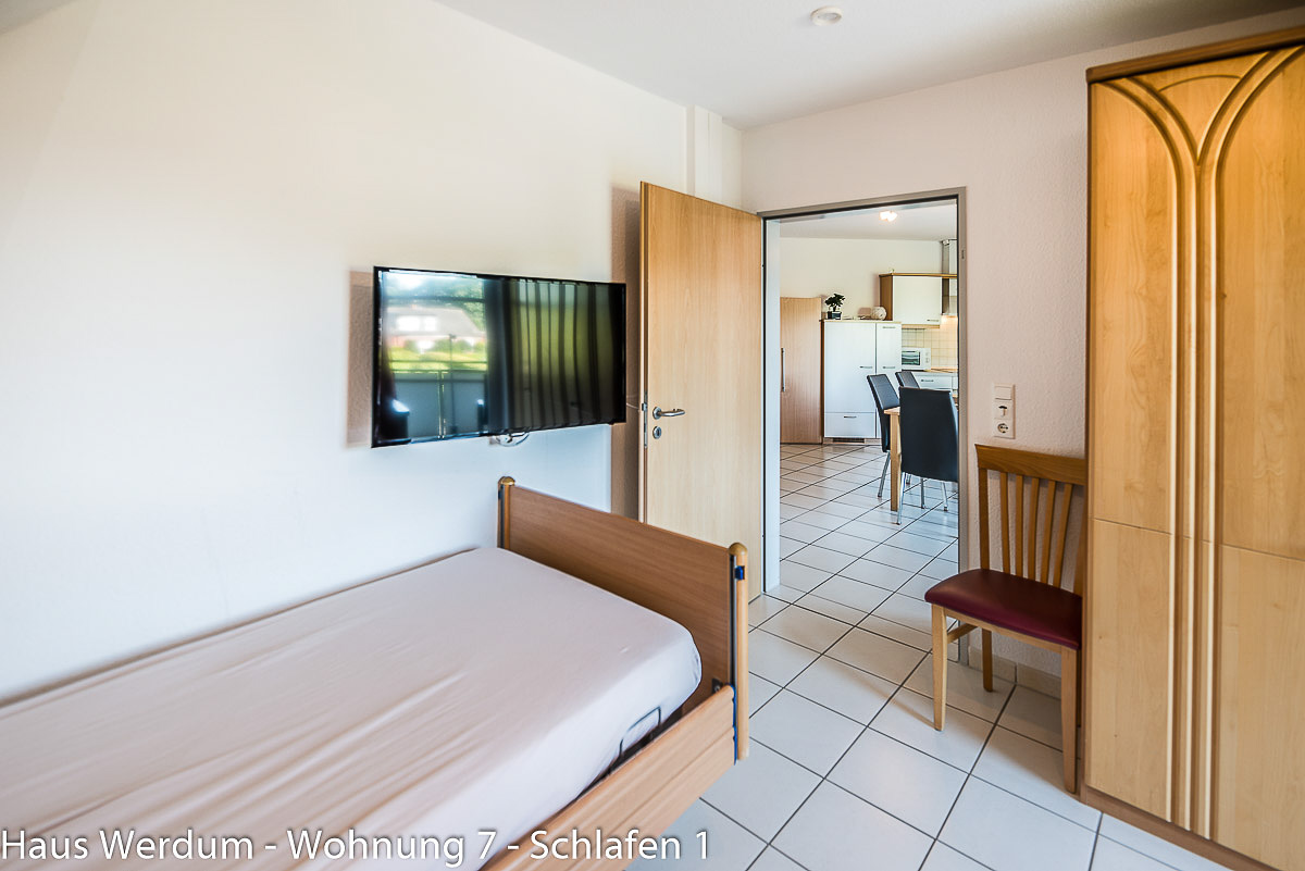 Blick vom höhenverstellbaren Pflegebett mit Seitengitter durch die geöffnete Tür in die Wohnküche. Der Zugang ist laut Reisen für Alle 93 cm breit und schwellenlos; vor dem Schrank sind 350 × 150 cm Bewegungsfläche dokumentiert.