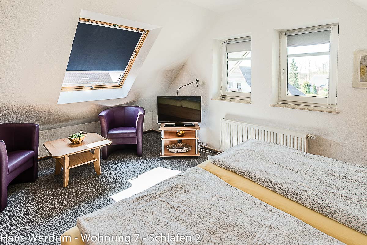 Schlafzimmer 2 der Wohnung 7 mit zwei Betten, TV, zwei Sesseln und Tisch unter Dachschrägen. Der Schlafraum liegt oberhalb der Wohnküche und ist nur über die Holztreppe erreichbar; ein barrierefreier Rollstuhlzugang besteht nicht.
