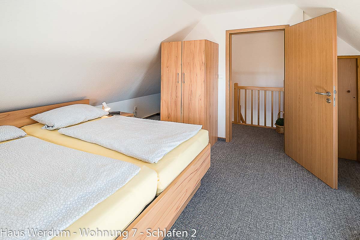 Schlafzimmer 2 der Wohnung 7 mit Betten, Kleiderschrank und geöffneter Tür zum Treppenbereich. Der sichtbare Ausgang führt zur Holztreppe; der Schlafraum ist deshalb nicht barrierefrei und für Rollstuhlfahrer nicht erreichbar.