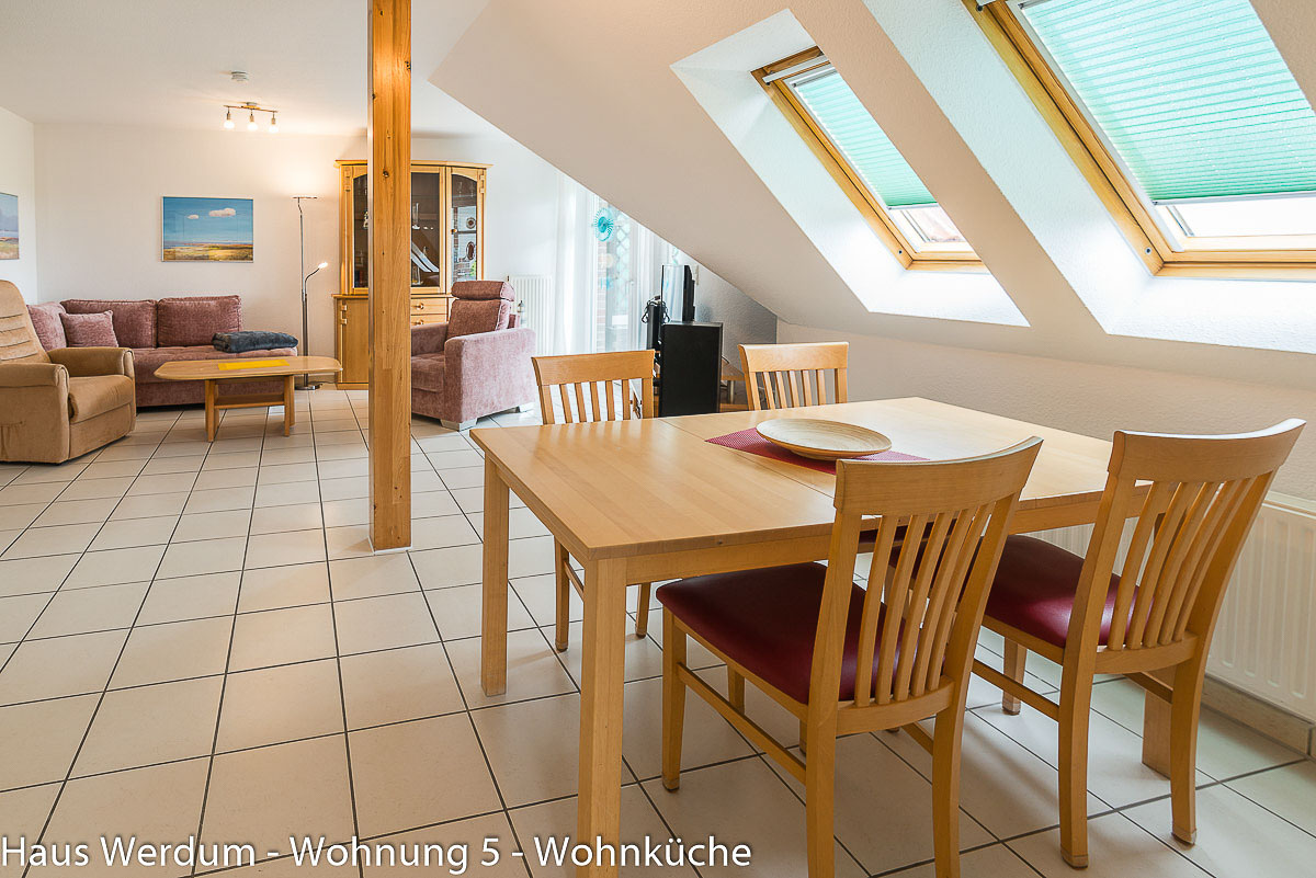 Heller, weitläufiger Essbereich in Wohnung 5 im Haus Werdum mit einem Holzesstisch, vier Holzstühlen mit roten Polstern und schwellenlosem Fliesenboden unter zwei großen Dachfenstern mit Plissees. Der offene Raum geht barrierefrei und ohne Stufen in den angrenzenden Wohnbereich mit Couchgarnitur und Sesseln über und bietet breite Fahrwege für Rollstühle