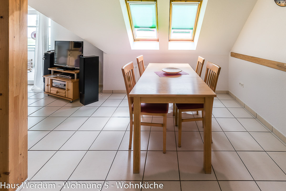 Heller Essbereich in Wohnung 5 im Haus Werdum mit langem Holzesstisch, vier gepolsterten Stühlen und schwellenlosem Fliesenboden unter zwei Dachfenstern. Im Hintergrund ist ein TV-Board aus Holz mit Fernseher und Lautsprechern neben dem stufenlosen Zugang zum Balkon zu sehen