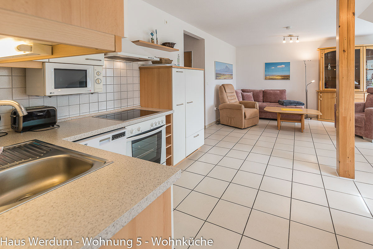 Helle Küchenzeile in Wohnung 5 im Haus Werdum mit Spüle, Cerankochfeld, Backofen, Mikrowelle und Kühlschrank. Der stufenlose Fliesenboden führt direkt in den angrenzenden Wohnbereich mit Sofa, Fernsehsessel und Couchtisch bei breiten Durchgängen