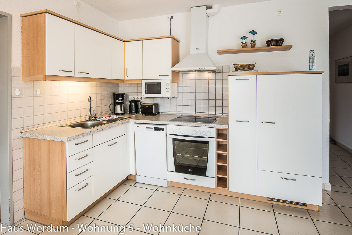 Einbauküche über Eck in Wohnung 5 im Haus Werdum mit hellen Fronten, integriertem Geschirrspüler, Backofen, Cerankochfeld, Dunstabzugshaube und Mikrowelle auf schwellenlosem Fliesenboden mit breiter Freifläche davor