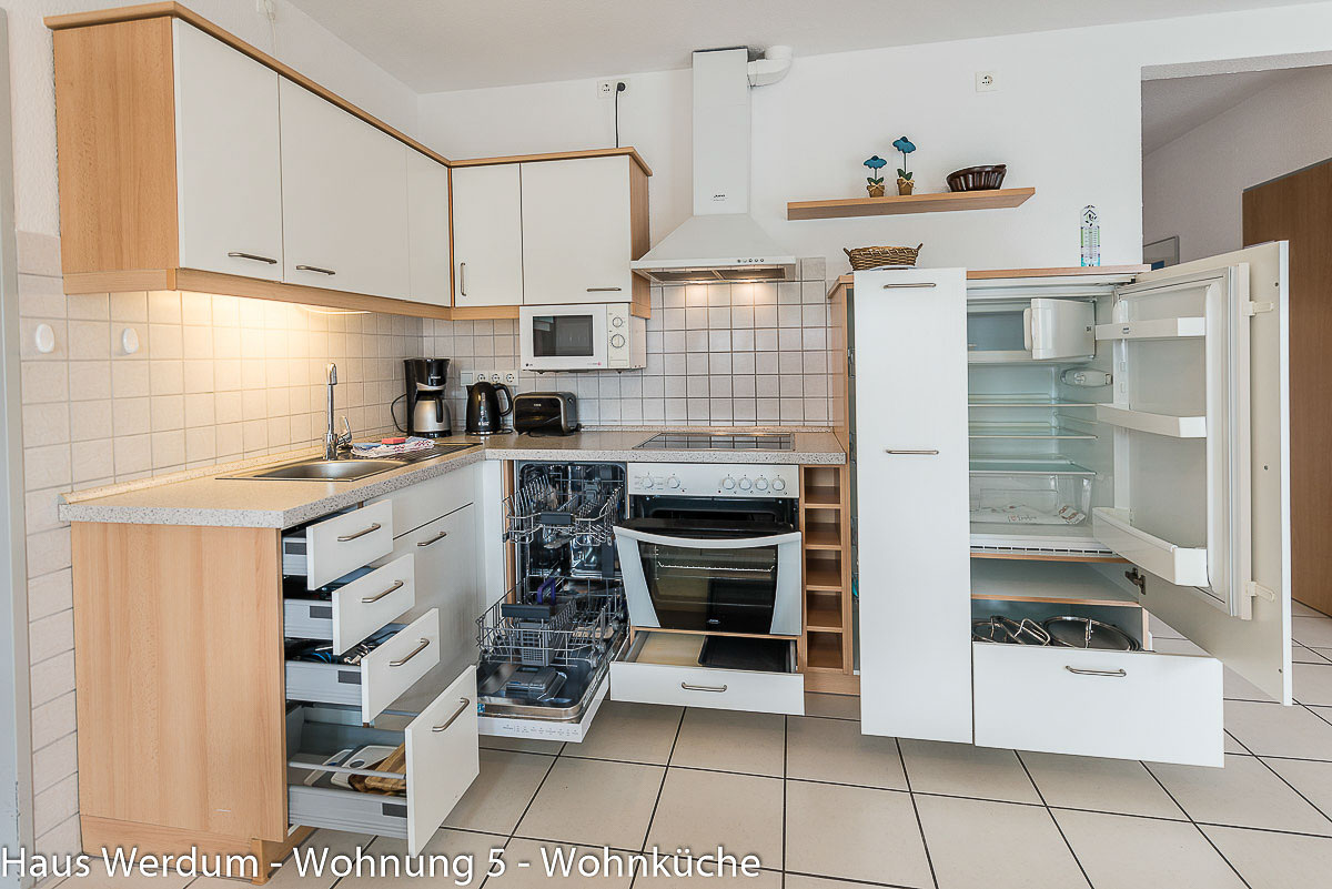 Geöffnete Küchenzeile in Wohnung 5 im Haus Werdum mit ausgelesenem Geschirrspüler, geöffnetem Kühlschrank, Backofen und weit ausgezogenen Schubladen auf schwellenlosem Fliesenboden zur Demonstration der Zugänglichkeit