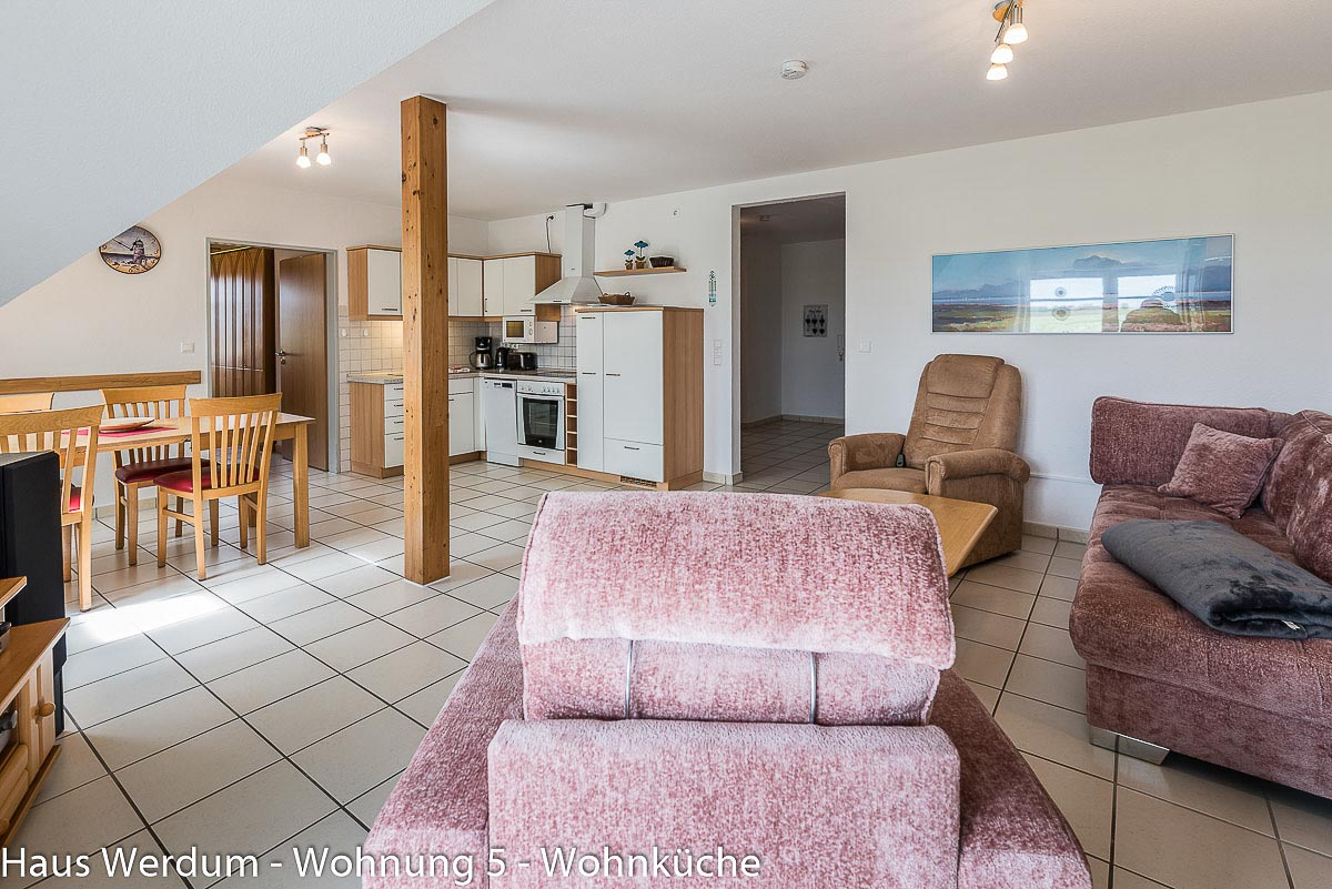 Offener Allraum in Wohnung 5 im Haus Werdum aus der Perspektive des Wohnzimmers mit Sofa, Sesseln, zentralem Holzpfeiler und freiem Blick auf die Küchenzeile, den Esstisch und den stufenlosen Fliesenboden“.