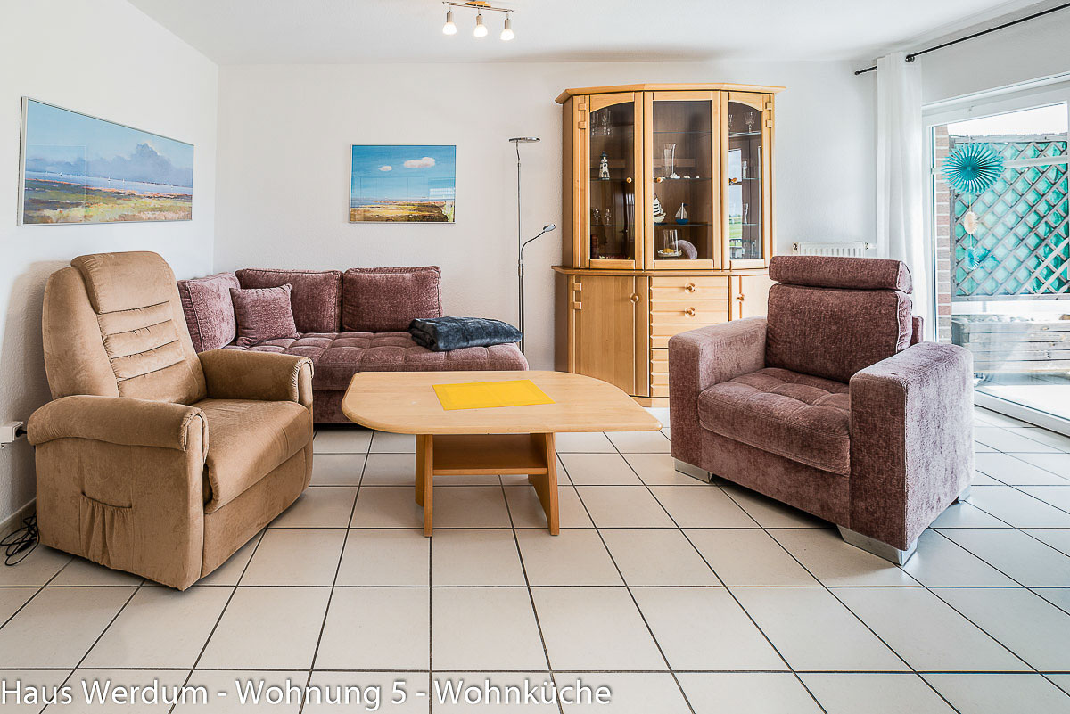 Helles Wohnzimmer in Wohnung 5 im Haus Werdum mit Eckcouch, zwei Sesseln und Holzvitrine auf schwellenlosem Fliesenboden. Rechts ist die große, barrierefreie Glasschiebetür zum überdachten Balkon geöffnet