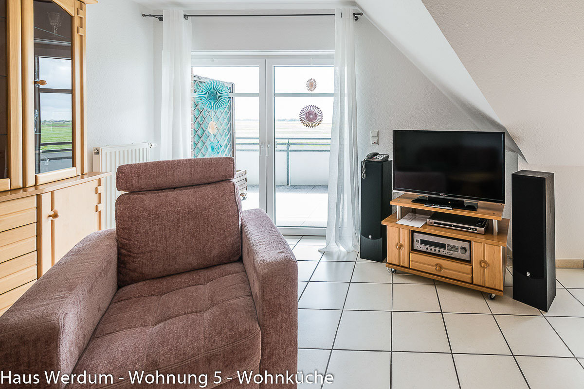 TV-Ecke in Wohnung 5 im Haus Werdum mit Flachbildfernseher auf fahrbarem Holz-Media-Board und großem Polstersessel auf schwellenlosem Fliesenboden. Im Hintergrund ist die geschlossene, barrierefreie Balkontür mit Weitblick ins Grüne sichtbar