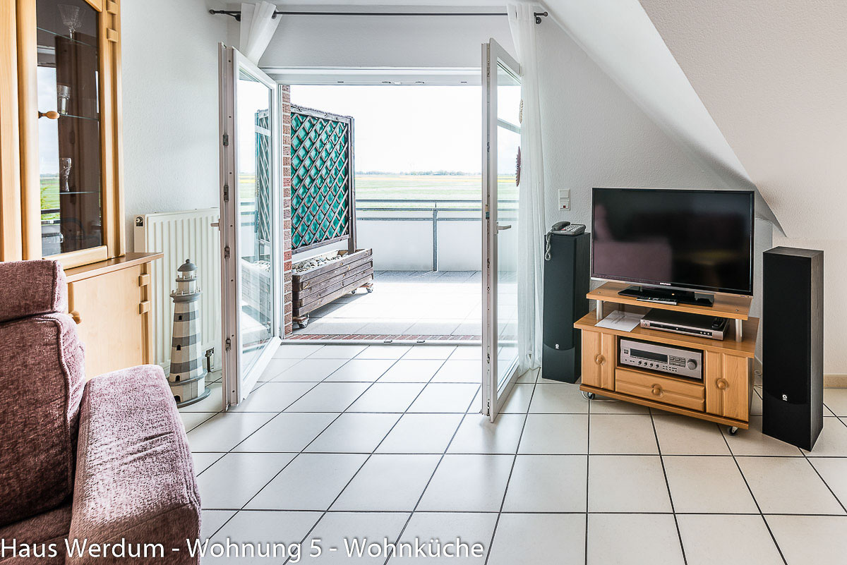 Weit geöffnete, zweiflügelige Balkontür in Wohnung 5 im Haus Werdum mit stufenlosem Übergang vom gefliesten Wohnbereich zum ebenen Plattenboden des Balkons. Rechts steht ein TV-Board mit Fernseher und links eine dekorative Leuchtturmsäule