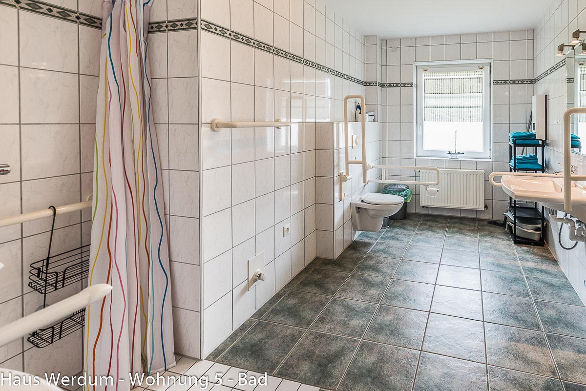 Großzügiges barrierefreies Badezimmer im Querformat mit Blick von der bodengleichen Dusche links auf den gesamten Raum. Mittig an der Wand befindet sich das wandhängende WC mit beidseitigen, weißen Stützklappgriffen. Rechts ist der unterfahrbare Waschtisch mit Spiegel angeordnet. Im Hintergrund befindet sich ein großes Fenster über einem flachen Heizkörper, und der Boden ist mit rutschhemmenden, graublauen Fliesen ausgelegt.