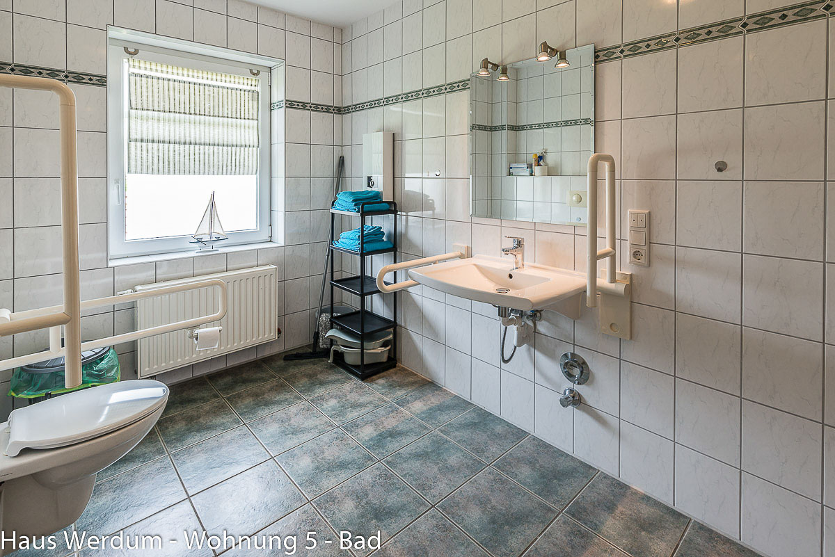 Barrierefreier Waschtisch im Querformat mit flacher Bauweise zur Unterfahrbarkeit für Rollstühle. Rechts und links am Waschbecken sind weiße Stützklappgriffe montiert. Darüber hängt ein großer Wandspiegel mit zwei Strahlern. Links im Hintergrund steht ein schwarzes Standregal mit blauen Handtüchern neben einem hellen Fenster mit Rollo. Am linken Bildrand ist das wandhängende WC mit Haltegriff angeschnitten.