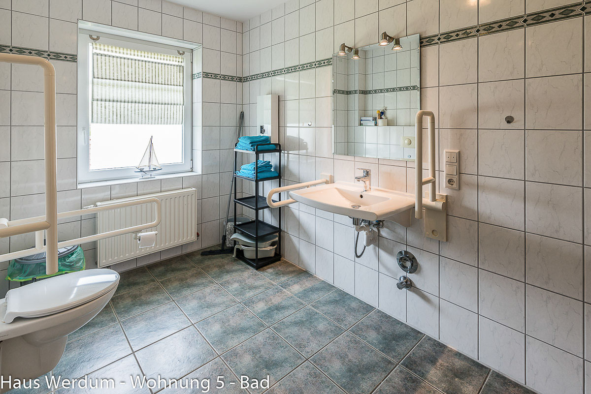 Barrierefreies Badezimmer mit unterfahrbarem Waschtisch und zwei stabil heruntergeklappten Stützgriffen. Über dem Waschbecken befindet sich ein Wandspiegel mit zwei Strahlern. Links im Hintergrund steht ein schwarzes Standregal mit blauen Handtüchern neben einem hellen Fenster mit Rollo. Am linken Bildrand ist das wandhängende WC mit klappbarem Haltegriff zu sehen.
