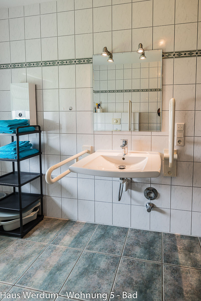 Barrierefreier, unterfahrbarer Waschtisch im Hochformat mit Einhand-Armatur und flachem Siphon. Der linke weiße Stützgriff ist heruntergeklappt, während der rechte Stützgriff platzsparend nach oben geklappt an der Wand ruht. Darüber hängt ein quadratischer Wandspiegel mit zwei Strahlern. Links steht ein schwarzes Metallregal mit blauen Handtüchern auf den blau-grau melierten Bodenfliesen.