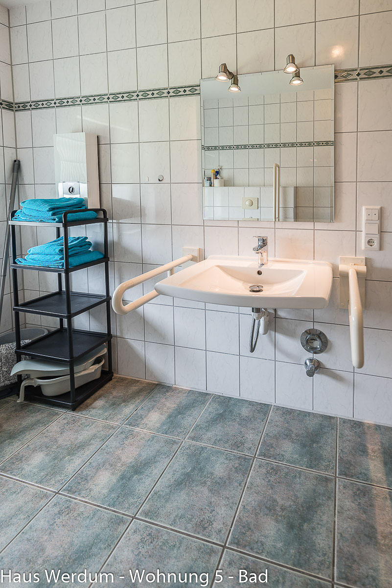 Barrierefreier, unterfahrbarer Waschtisch im Hochformat mit Einhand-Armatur und flachem Siphon. Beide weißen Stützgriffe an den Seiten sind stabil nach unten geklappt. Darüber befindet sich ein quadratischer Spiegel mit zwei Strahlern. Links steht ein schwarzes Standregal mit blauen Handtüchern auf den blau-grau melierten