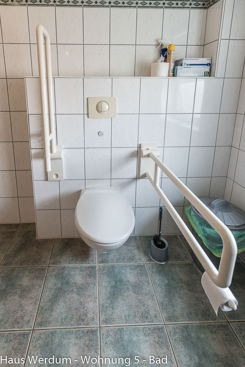 Barrierefreie Toilette mit einem links hochgeklappten und einem rechts heruntergeklappten Stützgriff inklusive integriertem Toilettenpapierhalter. Das wandhängende WC befindet sich vor einer halbhohen, weiß gefliesten Vorwandinstallation mit Spülungsknopf. Rechts daneben stehen eine Klobürste und ein Mülleimer, darüber befindet sich eine Ablagenische auf den blau-grau melierten Bodenfliesen.