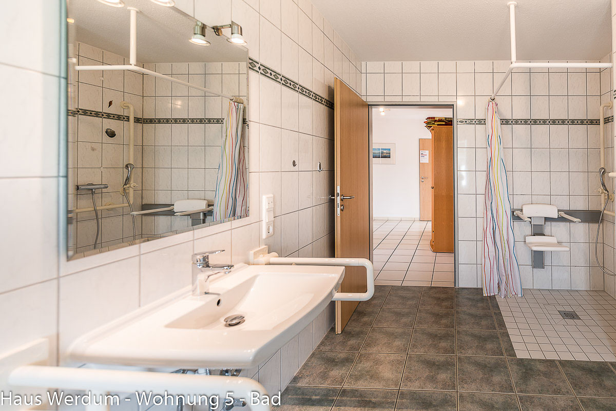 Barrierefreies Badezimmer mit Blick auf die ebenerdige Dusche inklusive an der Wand montiertem, klappbarem Duschsitz und Haltegriffen rechts. Im Vordergrund links das unterfahrbare Waschbecken mit klappbarem Stützgriff und großem Spiegel. Mittig führt eine breite, schwellenlose Holztür in den angrenzenden Wohnbereich mit hellem Fliesenboden.