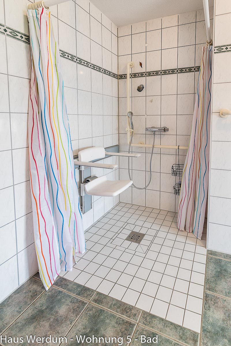 Bodengleiche, barrierefreie Dusche mit einem an der Wand montierten, weißen Duschsitz inklusive Armlehnen. Der Duschbereich ist schwellenlos mit kleinen rutschhemmenden weißen Fliesen ausgelegt und verfügt über einen mittigen Abfluss. An den Wänden befinden sich Haltegriffe, eine Duscharmatur und ein Duschvorhang mit bunten Längsstreifen, der an einer Eckschiene hängt.