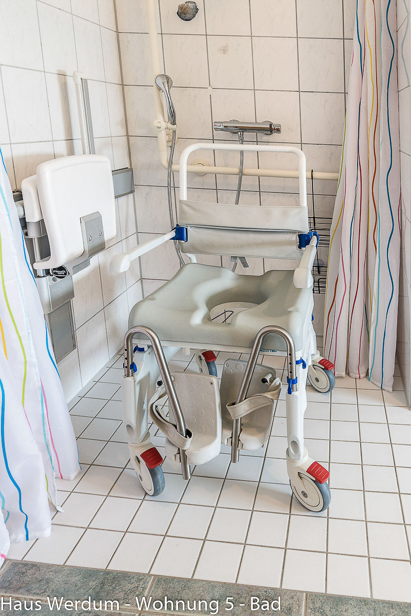 Mobiler Duschrollstuhl mit Feststellbremsen, Armlehnen und Fußstützen steht zentral in einer bodengleichen Dusche mit weißen Fliesen. Links im Hintergrund ist ein weißer Wand-Duschsitz hochgeklappt. An der Rückwand befinden sich eine Duscharmatur und waagerechte Haltegriffe. Ein bunt gestreifter Duschvorhang rahmt den schwellenlosen Duschbereich ein.