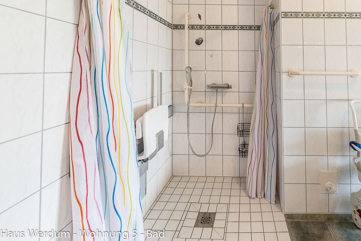 Bodengleiche, barrierefreie Dusche im Querformat mit schwellenlosem Einstieg und weißen Mosaikfliesen. An der linken Wand befindet sich ein hochgeklappter, weißer Wand-Duschsitz mit Armlehnen. Die Rückwand zeigt eine Duscharmatur mit Handbrause an einer Gleitstange und waagerechte Haltegriffe. Zwei bunt gestreifte Duschvorhänge flankieren den Duschbereich.