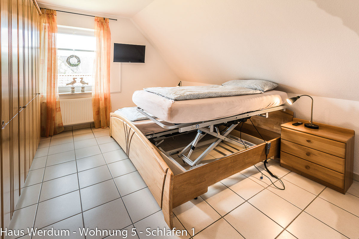 Barrierefreies Schlafzimmer im Haus Werdum Wohnung 5 mit hochgefahrenem, höhenverstellbarem Pflegebett. Sichtbare Scherenhebe-Konstruktion im Holzrahmen, Nachttisch, Fliesenboden und Fenster