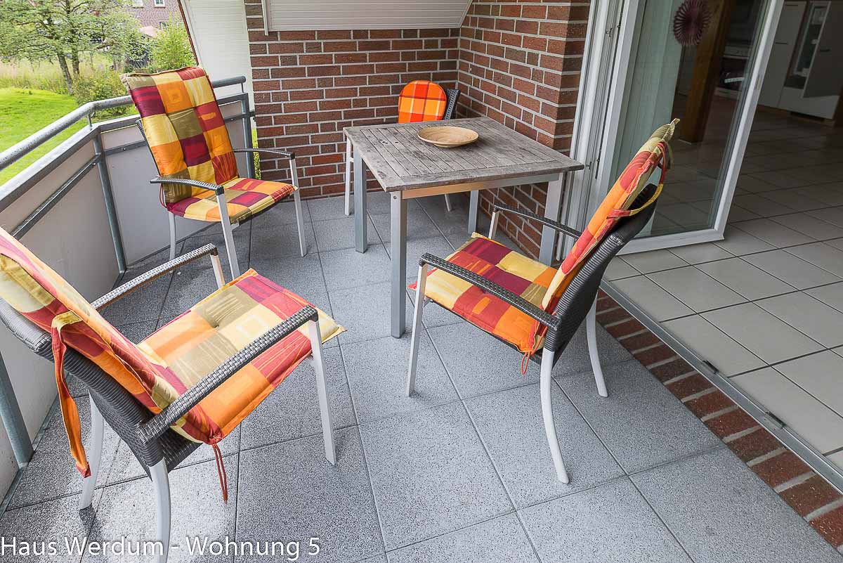 Balkon der Wohnung 5 im Haus Werdum mit grauen Steinplatten, quadratischem Holztisch und vier Gartenstühlen mit gemusterten Auflagen. Schwellenloser Übergang zum gefliesten Innenbereich erkennbar