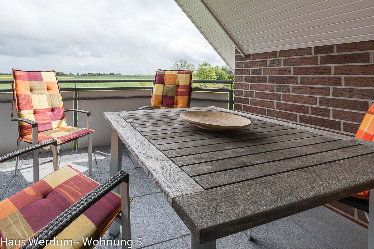 Perspektivische Ansicht des überdachten Balkons in Wohnung 5, Haus Werdum. Massiver Holztisch, Gartenstühle mit Auflagen und freier Ausblick auf die ostfriesische Landschaft